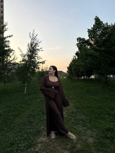 A woman stands in a grassy area in Odintsovo, Russia. She wears a brown dress with a matching long cardigan and light-colored sandals. The location is a grassy area with trees, possibly a park or green space. The sky is a pale gradient of blue and yellow, indicating either dawn or dusk. The presence of buildings suggests an urban setting, and based on the request, this urban setting is Odintsovo, Russia.