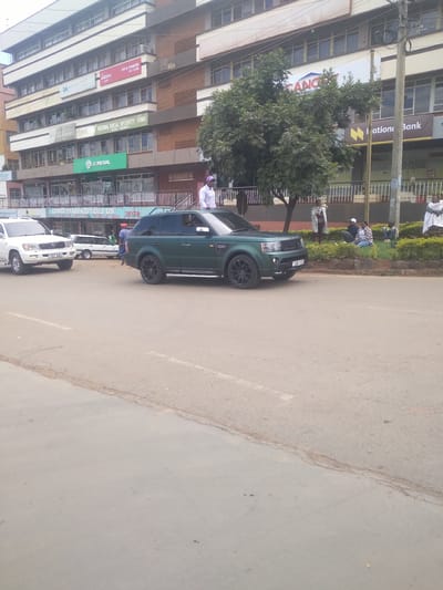 The image depicts a street scene in Nyeri, Kenya, featuring buildings with prominent signage.  Notable signs include "National Social Security Fund," "Old Mutual," "CENDCO PHARMACEUTICALS LTD.," "Jubilee" (LIFE & PENSION 2ND FLOOR), and "Iation Bank". Parked vehicles, including a dark green SUV and several white vehicles, are visible along the roadside. People are seen near the edge of the road. The road itself appears to be asphalt with some visible wear. A tree is also present.