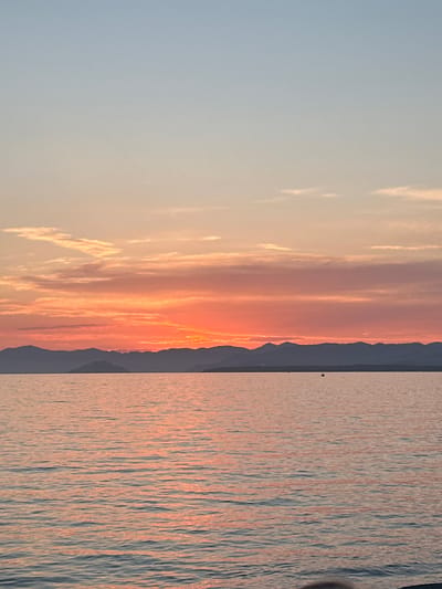 The image depicts a tranquil seascape at sunset, likely taken from a boat or the shore in Fethiye, Turkey. The sky transitions from a pale blue at the top to vibrant shades of orange and pink as it nears the horizon, indicating the setting sun. Wispy clouds are scattered across the sky, catching the colorful light. A silhouetted mountain range forms a dark line across the horizon, separating the sky from the sea. The water is calm, reflecting the warm hues of the sunset, with gentle ripples creating a shimmering effect. A small, dark object, possibly a buoy or a distant boat, is visible in the water. The overall atmosphere is serene and picturesque, capturing the natural beauty of a coastal sunset.