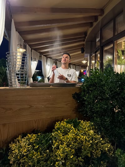 A man is standing behind a wooden counter, holding a white spray bottle and spraying mist into the air. He is wearing a white t-shirt with a small black tag. Stacked metal baskets are to his left, and plates are stacked on the counter in front of him. The scene appears to be an outdoor seating area of a restaurant or cafe, with white curtains and wooden beams overhead. Lush green and variegated foliage are visible in the foreground and background. The lighting suggests it is nighttime or dusk. The location is identified as Sirius, Russia.