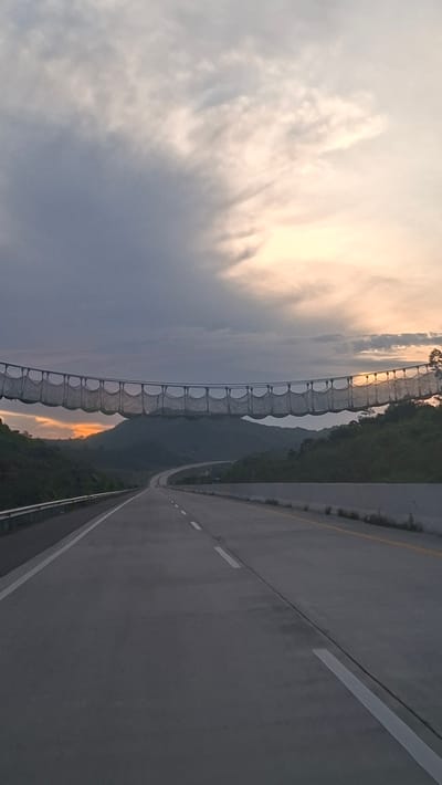 The image captures a highway passing through a mountainous landscape at dusk. A long, curved canopy structure, possibly a pedestrian bridge or a decorative element, spans across the highway in the mid-ground. The structure is composed of metallic framing with a mesh-like material forming individual suspended sections. The highway itself is paved and features lane markings. The sky above is filled with dramatic clouds, with the setting sun casting a warm glow on the horizon, visible between the mountains and through the canopy. The surrounding hills are covered in dense vegetation. The image appears to be taken from a moving vehicle, given the perspective. The location is identified as unknown, Indonesia.