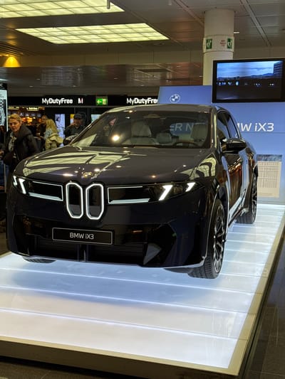 The image depicts a dark-colored BMW iX3 on display at an airport in Oberding, Germany. The car is positioned on a raised, illuminated platform. The front of the car, with its distinctive BMW grille and headlights, is facing the camera. In the background, there's a large sign that includes the words "MyDutyFree," suggesting the location is near duty-free shops. Passengers are visible in the background, and there's a screen and a post with "AED" written on it.