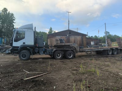 A white semi-truck with a large trailer is parked on a dirt lot in the city of Gremoochi, Russia. The truck is white with black accents and has large, rugged tires. The trailer is flatbed and carries two spare tires and a portion of a metal structure. In the background, there is a wooden building with a corrugated metal roof. Trees and power lines are visible behind the building. The sky is partly cloudy, with a mix of white clouds and blue sky. The ground is covered in dirt, with some scattered debris and sparse vegetation. The scene appears to be an industrial or storage area. There are no people visible in the image.