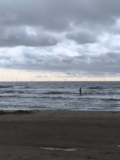 A solitary person stands in the choppy, grey sea, facing away from the camera. The water is dark and restless, with waves breaking around their knees. The beach in the foreground is dark and damp, with some small mounds of sand. In the distance, a vast wind farm stretches across the horizon, marked by numerous white turbine structures. The sky is overcast with heavy, dark grey clouds, with a pale sliver of lighter sky near the horizon. The scene evokes a sense of isolation and the power of nature. The image is likely captured on the coast near Bergen, Netherlands, given the presence of the offshore wind farm.