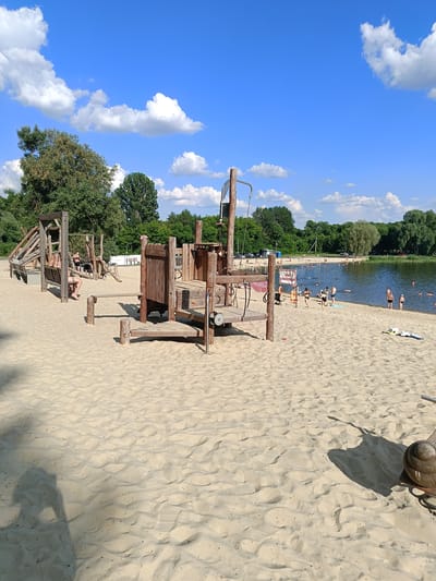 The image depicts a sunny day at a sandy beach adjacent to a body of water, likely a lake or river, bordered by a treeline. In the foreground, a wooden playground structure is situated on the sand. Several individuals are visible on the beach and in the water, some appearing to be swimming or relaxing. The sky is blue with scattered white clouds. The scene appears to be a public recreational area. The text on the sign in the middle of the beach reads "ПОСТ СПАСАТЕЛЯ," which translates to "Lifeguard Post" in Russian. This indicates the location is likely a designated swimming or bathing area. Based on the information provided, this is a public beach located in the city of Gubkin, Russia.