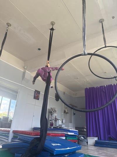 The image depicts an aerial silk performer suspended in the air, performing in what appears to be an indoor studio. A woman, wearing a purple and black patterned leotard, is in a split position, held up by black aerial silks. She is positioned below a large black hoop, which is also suspended from the ceiling by cables. The ceiling is made of white panels, and the walls are painted in light gray and white. To the right, there are stacks of blue and red mats, suggesting a gymnastics or dance studio environment. A purple curtain hangs on the right side of the image, and some shelving units with decorative items and a trophy are visible in the background. A framed picture is hanging on the wall. The lighting suggests it is daytime, likely indoors, with natural light coming from windows visible in the background. The overall scene conveys a sense of athleticism and artistry. There is no visible text in the image.