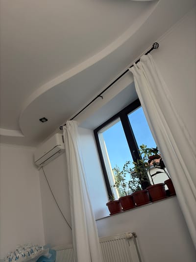 The image presents an interior scene, understood to be located in Topolovgrad, Bulgaria, captured from a low, upward-angled perspective. The room features white walls and a white ceiling. A distinctive architectural detail on the ceiling is a curved false ceiling section with a recessed, square-shaped light fixture, which is currently unilluminated. White decorative trim molding runs along the edge of this ceiling feature.
A dark, possibly black, metal curtain rod with a decorative caged-sphere finial on the right side is mounted above a window. White fabric curtains are drawn open, framing the window and allowing natural light to enter. The window itself has dark frames and reveals a clear blue sky outside, suggesting daytime. Green foliage from exterior trees is partially visible through the lower sections of the window.
On the white wall to the left of the window, a white 'Tesla' branded split-system air conditioning unit is mounted. A light gray cable or tube extends vertically downward from the unit along the wall. Along the interior window sill, five potted plants in uniform reddish-brown plastic pots are arranged. Below the window, a white panel radiator equipped with a thermostatic valve is partially visible. In the bottom-left corner of the image, a collection of clear plastic items, possibly cups or containers, adorned with blue butterfly patterns, rests upon a light blue surface or bag. No individuals are visible within the frame.