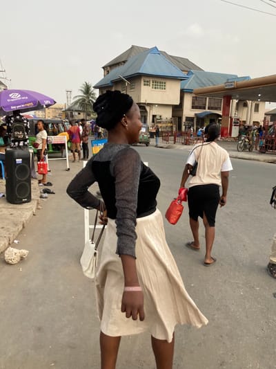 This image depicts a bustling street scene in Ura Nta Umuarandu, Nigeria.
In the foreground, a female with dark skin is captured in a dynamic pose, looking over her right shoulder towards the viewer's left. She wears a black head covering, a long-sleeved top featuring a black velvet bodice and shimmering dark grey sheer sleeves, and a flowing cream-colored pleated skirt that extends to just above her knees. A light pink wristband is visible on her right wrist. She holds a white quilted shoulder bag with a dark chain strap in her left hand. Her expression is a faint smile.
Immediately behind her, in the midground, another female with dark skin walks away from the camera, carrying a red bag in her right hand. She is dressed in a beige vest over a white short-sleeved top, a black pencil skirt, and dark flip-flops.
The background reveals a busy urban environment. To the left, a purple "PalmPay" branded umbrella shades a street vendor's stall, displaying items including a black backpack with "STAR BOY" text. A large black speaker rests on the ground nearby. A green motorized tricycle (Keke Napep) is partially visible behind the stall. Prominently in the center background stands a multi-story building with light-colored walls and a distinctive blue, pyramidal-shaped roof. To its right, another building, possibly a commercial establishment, features a red fuel pump structure with "BICHAZWILL" visible on an awning, suggesting a gas station. Numerous other pedestrians are scattered throughout the background, engaged in various activities on the street and near the buildings. The sky is a uniform, pale grey, indicating an overcast day or atmospheric haze. The ground is paved, with a concrete curb visible on the left side featuring some debris.