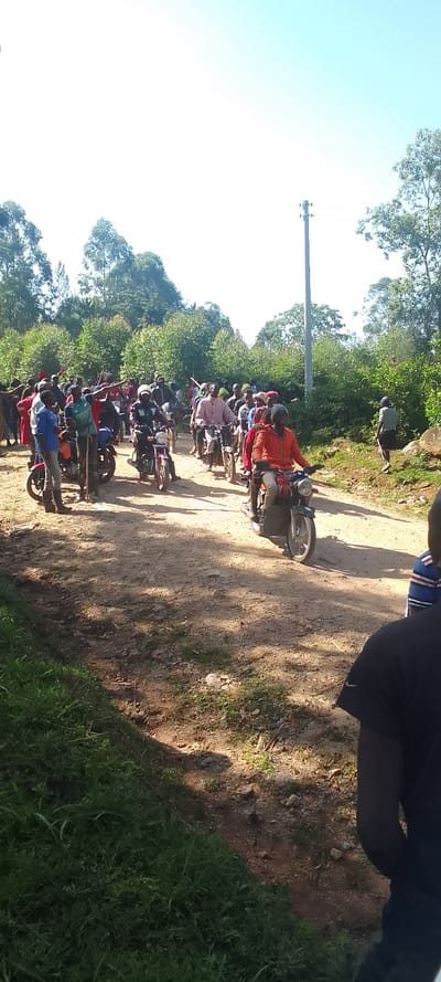 The image shows a group of people riding motorcycles on a dirt road in Isukha ICHINA, Kenya. The coordinates provided place the scene in a rural area outside of the town of Isukha. The group of people is mostly male, wearing casual clothing. They are riding motorcycles in a line, with some carrying passengers. The road is dusty and dry. The surrounding environment is forested, with trees and brush on both sides of the road. There is a utility pole in the background and a person walking on the right side of the road.