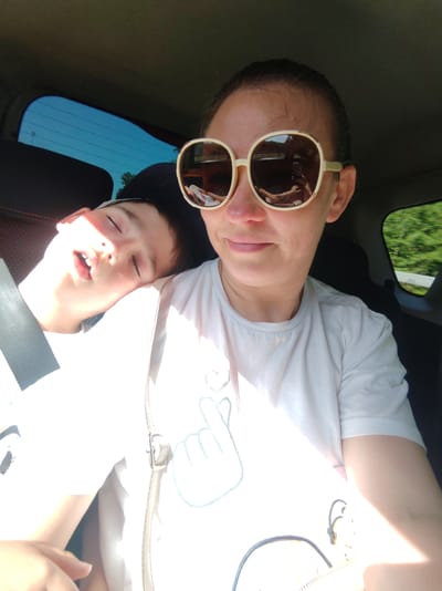 In a car, a woman with sunglasses and a white t-shirt is taking a selfie. Next to her, a young boy is sleeping, his head resting on her shoulder. The car is moving, as indicated by the blurred scenery outside the windows. The overall scene suggests a family road trip on a sunny day, possibly in or around Sosnovka, Russia, a town known for its forested surroundings.