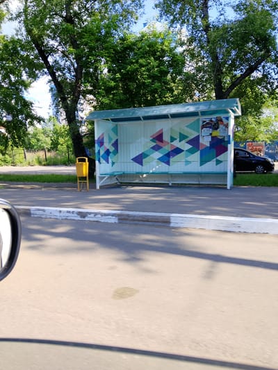 A black sedan is parked at a bus stop on a sunny day in Abakan, Russia. The bus stop shelter has a translucent green roof and is decorated with colorful triangular patterns. A yellow trash can stands to the left of the bus stop. The scene is viewed from a moving vehicle, as indicated by the reflection of a side mirror in the lower left corner of the image.