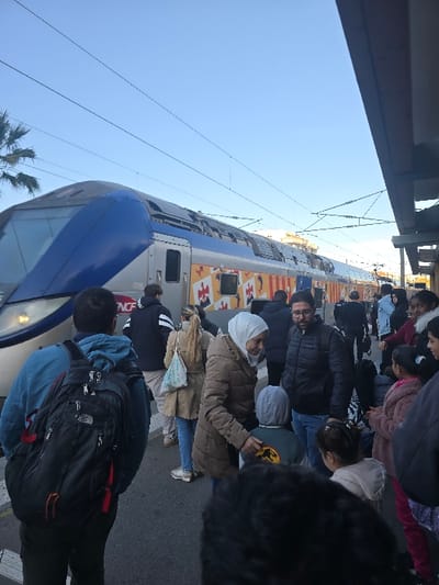 A crowd of people surrounds a blue and silver train, likely boarding or disembarking. Many wear coats, suggesting a cool climate. Backpacks and bags are visible. The train features a painted design and bears the SNCF logo. Overhead power lines stretch above the train, and part of a building structure is visible at the top left.