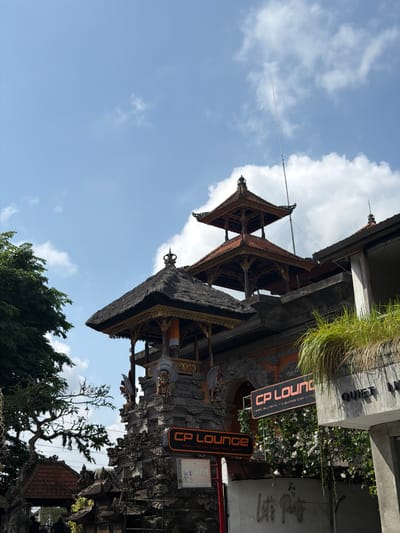 The image shows a sunny day in Ubud District, Indonesia, with a clear blue sky dotted with fluffy white clouds. In the foreground, an ornate Balinese gate stands prominently, featuring intricate stone carvings and traditional tiered roofs. The gate is adorned with statues of mythical creatures and decorative gold accents.
A prominent orange sign with white lettering reads "CP LOUNGE restaurant, tapas bar, nig". Below it, another orange sign with white lettering says "CP LOUNGE drink, dance, swim". To the right of the gate, a modern concrete building displays a sign that reads "QUIET IN". Lush green vegetation, including grass and vines with small white flowers, cascades down from the building and around the lounge signs.
To the left of the gate, a large, leafy green tree provides a natural contrast to the man-made structures. A thin, tall antenna or pole extends vertically into the sky from behind the gate's roof. In the lower part of the image, two individuals are partially visible, walking towards the gate. An ATM sign is also visible on a post near the gate. The overall scene captures a blend of traditional Balinese architecture and modern commercial establishments in a tropical setting.