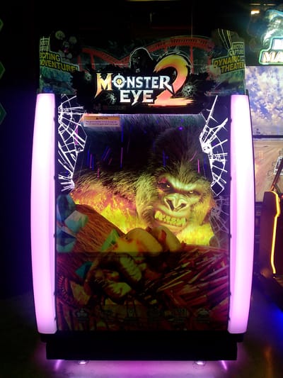 The image shows an arcade game machine with a large gorilla graphic and the title "MONSTER EYE 2". The machine has a cracked screen effect overlaying the graphics. It is lit with purple neon lights on the sides. The machine is located in a dimly lit arcade, with other game machines visible in the background, including one with a racing theme. The bottom of the machine has icons for "Shaking Seat", "Air Blow", "Motion Capture", "Surround Sound", "Full HD 1080P", and "Atmosphere Lighting". There is a caution sign on the machine that reads "This game machine moves during use. Do not touch the game when it is in use." No people are visible in the image. The location is not specified in the image or the provided crops.