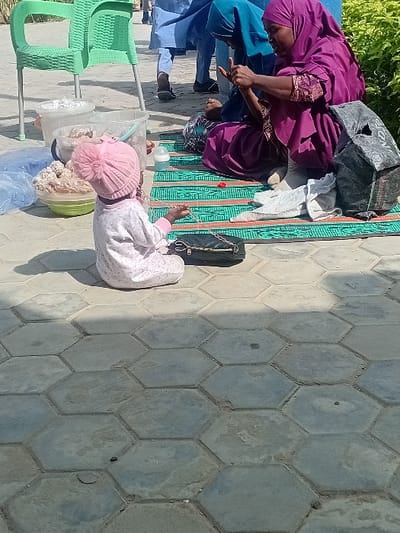 The image depicts an outdoor daytime scene in the city of Kwazailewa, Nigeria, set on a surface of hexagonal concrete pavers. In the foreground, a young child, dressed in a light-colored long-sleeved outfit and a pink knitted hat with a pom-pom, is seated on the pavers, facing away from the camera. The child holds a small, dark object in one hand and is positioned beside a small black purse with a chain strap.
To the child's right, two women are seated on a green mat featuring a repeating pattern. The prominent woman wears a deep purple hijab and matching full-length attire. Her hands are raised and engaged in a focused activity. Her lower legs and feet, covered in light-colored socks or fabric, are visible. Partially obscured behind her is a second woman, dressed in a teal hijab and dark clothing, also looking downwards.
Surrounding the individuals are various objects: an empty light green plastic chair is positioned to the left. Near it are two transparent plastic buckets, one containing white, fluffy material and the other with less distinct contents. A green bowl filled with numerous small, light-brown spherical items is on the ground. Several clear plastic bags are also visible. A baby bottle with a white top rests on the patterned mat. A crumpled black plastic bag is situated near the woman in purple.
In the background, several blurred figures are standing, one of whom wears a light blue traditional garment. A dense green hedge or bush lines the right side of the frame. The scene is under bright natural sunlight, creating clear shadows on the paved ground.