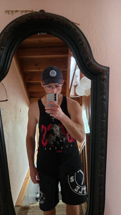 A person is taking a mirror selfie in what appears to be a home. The person is wearing a black baseball cap with a circular logo that includes the letters "VW" in white. They are also wearing a black tank top with a graphic print in red and white, and black shorts. The person is holding a smartphone, presumably to take the picture.
The mirror is large and ornate, with a dark, carved frame. It is reflecting the interior of the room, which includes wooden stairs and what appear to be white balloons hanging from the ceiling. The lighting suggests it is daytime. There is some visible text on the person's clothing. The tank top has a large red number "23" and other text, possibly a graphic or band name. The shorts have text in a circular design on the right leg. The overall scene is casual and appears to be an indoor setting. The reflection shows a light-colored wall and possibly a wooden floor.