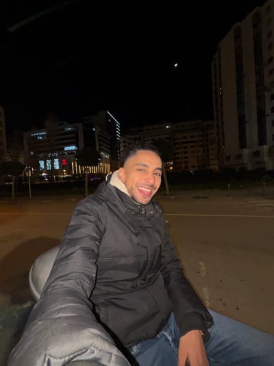 A smiling man with short hair and a black jacket poses at night. Behind him, illuminated buildings rise in the background, contrasting with the dark sky where a crescent moon is visible. The man appears to be sitting, with a person beside him wearing blue jeans. The setting suggests an urban environment at nighttime.