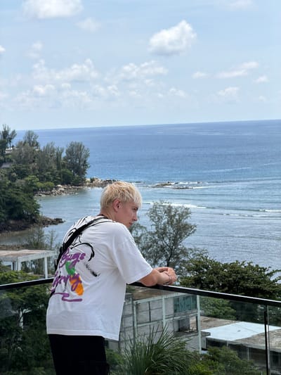 In Kamala, Thailand, a person with blond hair stands on a balcony overlooking the ocean. They are wearing a white t-shirt with a colorful abstract design on the back and black pants. Their arms are resting on the balcony railing, and they are looking out at the water. The ocean is a deep blue, and waves are gently rolling to shore. The coastline is lined with lush green trees, and in the distance, there are some buildings. The sky is partly cloudy with a mix of blue and white.