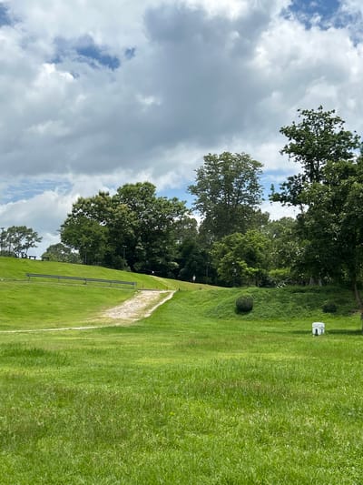 A wide-angle, eye-level shot captures a vibrant green grassy landscape under a partly cloudy sky in Chiang Mai City Municipality, Thailand. A light-colored gravel path winds its way up a gentle slope on the left side of the frame, bordered by lush green grass. A metal railing follows the contour of the slope. Several trees of varying sizes and foliage density dot the midground and background, creating a sense of depth. Some trees are tall and broad, while others are more slender. A few benches are visible on the upper left portion of the slope, suggesting a park or recreational area. In the mid-right, a small, white decorative structure with a pagoda-like roof sits in the grass. Two rounded bushes are also present in the midground. The foreground is dominated by a dense expanse of green grass, with some variations in texture and color. The sky is a mix of bright blue patches and large, fluffy white and gray clouds.