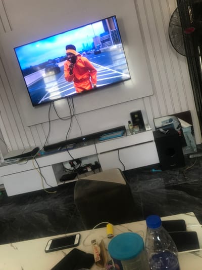 The image depicts an indoor living space in Akure, Nigeria. A large, wall-mounted flat-screen television prominently displays a male figure wearing an orange hooded tracksuit and dark headwear on what appears to be an elevated urban roadway; he gestures with his right index finger to his mouth. Below the television, a white entertainment console holds a soundbar, a video game console, and various electronic devices with visible cables. On the right side of the console, a '5G' labeled object, small bottles, and a blue tablet are visible. The room has white walls, some with vertical stripes, and a dark, glossy, marbled tile floor. A black stand fan is partially visible on the far right. A framed image of a person's legs leans on the floor. In the foreground, a white table features two smartphones, a plastic water bottle with a blue cap, a yellow lighter, and other small objects. A dark and light-toned ottoman sits in the mid-ground.