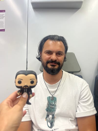 A man with dark hair and a beard, wearing a white t-shirt and a necklace with a pendant, is holding a Funko Pop figure in his left hand. The figure depicts a man in a suit with dark hair and a beard. The man is seated in a chair in front of a white wall. To the left of the man, a portion of a poster with text in Catalan and English is visible. The text mentions "Renfe," indicating the location is likely on a Renfe train or at a Renfe station. Given the context and visual cues, the scene is likely within a train carriage of Renfe, a Spanish state-owned railway company, possibly in or near Sant Adrià de Besòs, Spain, which is served by Renfe's commuter rail lines. The lighting is bright and even, suggesting an indoor environment.