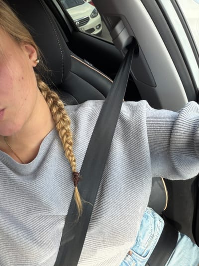 In a high-angle shot, a person is seen wearing a light gray turtleneck sweater and light-wash jeans, secured by a black seatbelt. A blonde braid rests on the sweater, close to the collarbone. The inside of a car is visible, with a white car reflected in the side mirror. Black leather car seats are partially visible.