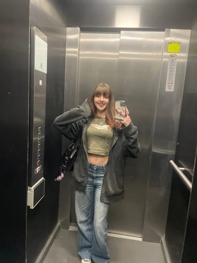 A young woman poses in an elevator, capturing her reflection in the mirror. She's wearing a green crop top, jeans, and a dark jacket, holding her phone up for the picture. The elevator has a control panel with illuminated buttons and a display screen. The overall atmosphere seems modern and confined.