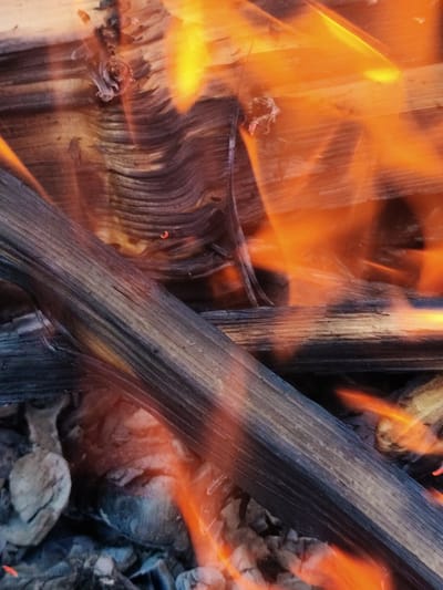 The image shows a close-up of a bonfire with burning logs and embers. The fire is producing bright orange and yellow flames, which are casting a warm glow on the wood. The wood itself is charred and appears to be dry, suggesting it is suitable for burning. The texture of the wood grain is visible, and some pieces are splintered. The overall scene depicts a natural and intense process of combustion. The location of this scene is in Радуга Снт (ботово), Russia.