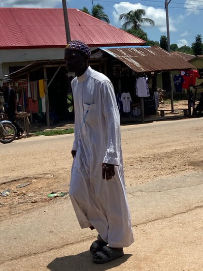 The image shows a man walking on a dirt road in Mararaba, Nigeria. The man is wearing a white robe and a patterned cap. He is walking towards the right side of the frame. In the background, there are buildings with red and brown corrugated metal roofs. Some of these buildings appear to be market stalls with clothing displayed for sale. A motorcycle is parked on the left side of the road. The sky is blue with some clouds, and palm trees are visible in the distance. The ground is a mix of dirt and some scattered debris.