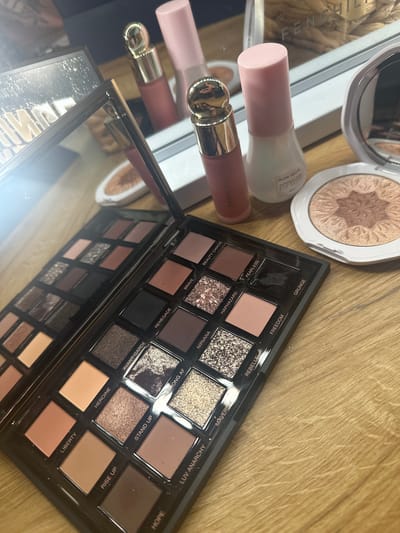 The image displays a collection of makeup products arranged on a wooden surface. In the foreground, an open eyeshadow palette is prominently featured, revealing multiple shades of eyeshadow. To the right of the palette, a compact highlighter is visible. In the background, two pink-capped bottles and a pink-hued lipstick are arranged. The scene appears to be indoors, with reflections visible in a mirrored surface behind the makeup. The lighting suggests a bright, well-lit environment. There is no indication of the city of Shumen, Bulgaria, or any specific location.