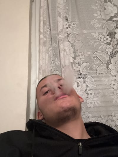 A man is exhaling a plume of smoke. He is wearing a black hooded jacket. Behind him is a window covered with a floral lace curtain. The wall below the window is light beige. The location is not discernible from the image and there is no indication it is in Goritsa, Bulgaria.