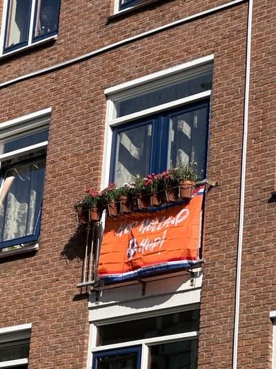 This is a medium-shot, eye-level, outdoor photograph taken on a sunny day, likely in the afternoon, judging by the harsh shadows. The scene is a brick-faced apartment building in The Hague, Netherlands. The perspective is slightly angled, looking upwards at a section of the facade.
The main subject is an orange banner with white lettering hanging from a window. The banner is decorated with a checkered pattern that resembles a football or sports logo. The text on the banner appears to be in white cursive script and reads "Hup Holland Hup!". This is a popular Dutch cheer associated with the national football team.
Below the banner, a window box is filled with lush green plants and small pink and red flowers. The window itself has a dark blue frame and white trim. Adjacent to this window are other windows of the building, also with dark blue frames and white trim, some of which offer glimpses into the interiors, revealing floral patterned curtains or other furnishings.
The building's exterior is constructed of red brick, laid in a stretcher bond pattern. Thin white horizontal bands run across the facade, possibly indicating different floor levels or architectural features. A metal downspout or pipe is visible near the banner. The overall impression is a typical urban residential scene, likely in The Netherlands, celebrating a sporting event or national pride. There are no people visible in the frame.