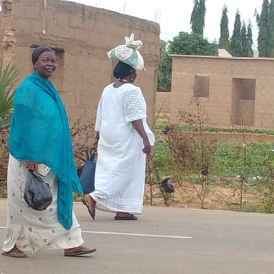 In Tempure, Nigeria, a woman is depicted walking on an asphalt road, wearing a white dress and carrying a large bag of goods on her head. She is accompanied by another woman who is walking beside her, wearing a blue headscarf and holding a black bag in her left hand. The background features a partially constructed building made of rough-hewn bricks, with incomplete window openings. Tall, slender trees are visible in the distance, along with dense green vegetation. The scene suggests a typical day in a Nigerian town, with people engaged in daily activities of commerce and transport.