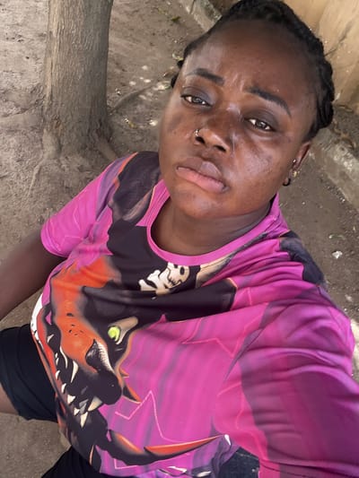 A close-up, slightly tilted selfie captures a young adult individual, likely female, with dark skin and dark braided hair. She wears a vibrant short-sleeved t-shirt predominantly in shades of pink and purple, featuring a bold graphic design on the chest. The design depicts a stylized animal head, possibly a dragon or mythical creature, with bright green eyes, orange and black facial features, and prominent teeth or horns. She also wears black shorts or leggings.
The individual has a neutral to slightly serious expression, with a nose piercing visible on her left nostril and an earring in her left ear. The setting is outdoors, under natural daylight, suggesting it is daytime. The background consists of a textured, unpaved ground or dirt path, with a thick tree trunk or large branch running horizontally in the lower left. The overall scene provides a glimpse of an outdoor environment in Kubwa, Nigeria. There is no visible text in the image.