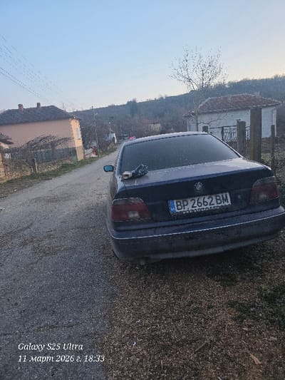 An undated image, captured by a Galaxy S25 Ultra at 18:33 on March 11, 2026, depicts a rural street scene in Dolna Beshovitsa, Bulgaria. A dark blue BMW E39 sedan, bearing Bulgarian license plate "BP 2662 BM," is parked angled towards the right side of a narrow, unpaved road. The vehicle occupies a portion of the dirt shoulder and the road itself. A pair of dark-colored gloves and a small metallic object rest on the car's trunk lid near the rear window.
To the left of the road, a two-story house with a reddish-brown tiled roof and light-colored walls stands, partially obscured by bare trees and utility lines. Further down the road on the right, a smaller white house with a similar tiled roof is visible. A prominent, bare deciduous tree is situated between the parked car and this smaller house. In the background, a tree-covered hillside rises against a clear, pale blue sky indicative of late afternoon or early evening. No individuals or active interactions are present in the scene. The ground is a mix of gravel, dirt, and sparse dry vegetation.