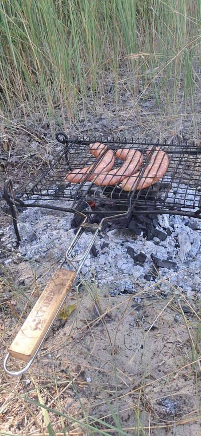 The image shows a close-up of a portable grill with several sausages cooking on it, placed over glowing coals. The grill has a metal frame and a wooden handle with the word "GRILL" visible. The surrounding area is covered in dry grass, sand, and ash from the fire. In the background, tall green and dry grass grows densely. The scene appears to be outdoors, possibly in a park or natural area, and given the context of Horishni Plavni, Ukraine, it suggests a recreational activity like a picnic or camping. There are no people visible in the frame.
