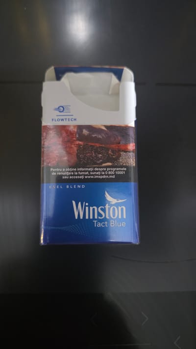 The image shows an opened pack of Winston cigarettes, specifically "Tact Blue" flavor, placed on a dark, reflective surface. The pack is mostly blue with white accents and features an image of tobacco leaves on one side. Visible text on the pack includes "FLOWTECH," "Winston," "Tact Blue," and "EXEL BLEND." There is also text in Romanian providing information about smoking cessation programs: "Pentru a obţine informaţii despre programele de renunţare la fumat, sunaţi la 0 800 10001 sau accesaţi www.imspdrn.md" which translates to "To obtain information about smoking cessation programs, call 0 800 10001 or visit www.imspdrn.md." The scene appears to be indoors, with soft lighting. The background is dark and slightly out of focus, with some subtle reflections and a faint vertical line, possibly indicating a window or a seam in the surface. There are no people or animals visible in the image. The focus is entirely on the cigarette pack, suggesting it is the main subject of the photograph. The overall impression is that of a product shot, potentially for an advertisement or inventory purpose. The location context of Chișinău, Moldova is indicated by the ".md" domain in the website address.