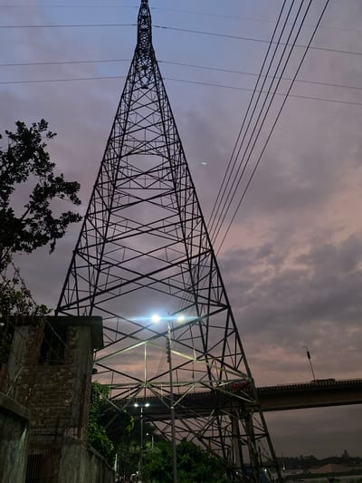 A low-angle, full-shot photograph captures a large, lattice-style electricity pylon dominating the frame against a twilight sky. The pylon, made of dark metal beams, tapers to a point at the top, with several high-voltage power lines strung horizontally across the sky to its right. To the left, the silhouette of a leafy tree partially obscures the sky and the pylon. In the foreground, a section of a brick structure, possibly a guardhouse or utility building, is visible. This structure has barbed wire fencing and a barred opening. Below and behind the pylon, a bridge is discernible, with a few indistinct figures and lights suggesting traffic or activity on it. The sky is filled with dramatic, dark clouds tinged with hues of purple and gray, illuminated by the fading light of dusk. The scene appears to be set in an industrial or semi-urban area, with a hint of vegetation and a body of water or canal visible in the distance. The overall mood is somber and somewhat industrial.