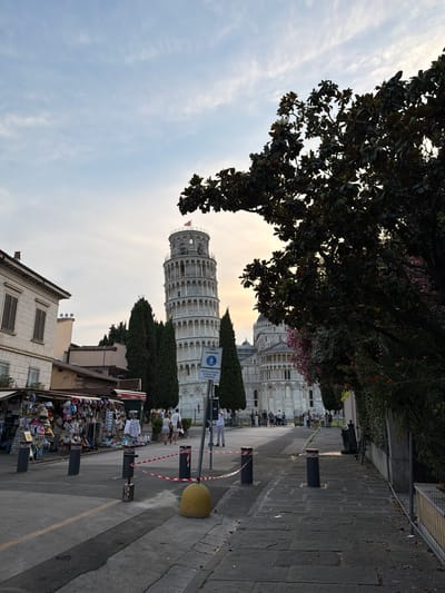The image captures a view of the Leaning Tower of Pisa and the surrounding square. The tower, a prominent white cylindrical bell tower, stands in the background, slightly tilted to the right. Adjacent to it is a larger white building with arched windows and a dome. In the foreground, a street is lined with souvenir shops displaying hats, clothing, and other tourist merchandise. A red and white barrier tape is strung between bollards, cordoning off a section of the walkway. Several people are visible in the scene, some walking along the street, others browsing the shops, and a few standing near the buildings. The sky above is a pale blue with scattered clouds, and the overall lighting suggests it is either early morning or late afternoon. Trees, some with dark green foliage and others with pink flowers, frame the scene. The ground is paved with cobblestones in some areas and asphalt in others. A sign with a pedestrian symbol and Italian text is placed near the tower.