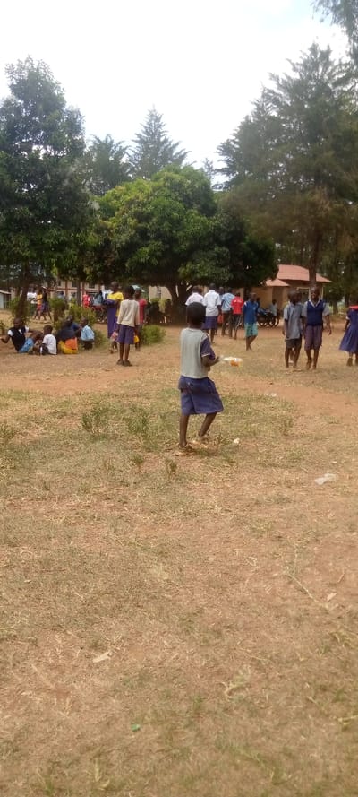 The image shows a group of children, mostly girls, in a grassy field in Malava, Kenya. The coordinates, latitude: 0.53, longitude: 34.811, correspond to a location near Malava town. There are around twenty children in the image, all wearing casual clothes. The children appear to be in their late elementary or early secondary school years. They are standing and walking in a small clearing in the field. There are trees and a building in the background. The grass is dry and brown, indicating a dry season. The children seem to be moving in a casual and relaxed way, perhaps after a school break or a play session.
