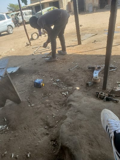 The image depicts an outdoor, unpaved workshop or informal mechanic area located in Kaduna South, Nigeria, under bright daylight conditions.
In the mid-ground, a male individual, appearing to be a young adult, is bent at the waist, facing slightly to the left, engaged in manipulating multiple metal components on the dusty, light brown ground. He wears a dark short-sleeved shirt, dark trousers, and light-colored sneakers. Scattered around him on the ground are various metal parts, including what appears to be a silvery automotive control arm, several wrenches, metal bars, and small metallic fragments. A small blue and yellow cylindrical container lies on its side to his left. Two weathered, dark-colored vertical poles, possibly supporting a makeshift overhead structure, are positioned to the right of the individual.
In the lower right foreground, a partial view of a left foot, encased in a white sneaker with black stripes, is visible, indicating the camera's close proximity to an observer. The ground directly in front of the camera is uneven and deeply rutted in places.
In the background, an unpaved open area or street features several vehicles, including a silver SUV, a white panel van, and a motorcycle, parked or idling. Behind the vehicles, a row of single-story buildings with worn, light-colored facades line the street. The sky above is clear and light blue.
