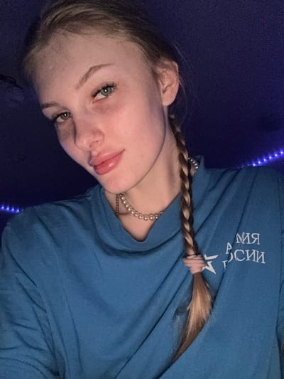 The image shows a young person, likely female, with fair skin, light-colored eyes, and light brown hair. Her hair is styled in a single braid, secured near the shoulder with a pink elastic, with the ends flowing freely. She is wearing a blue long-sleeved shirt with white Cyrillic text printed on it. Due to the image's orientation and possible reversal, the text appears as "ИИССО ЯМ АЯ" which, when read correctly and oriented, spells "МАЯ РОССИИ" (May Russia or Of Russia). She is also adorned with a pearl necklace and what appears to be a thin gold-toned chain beneath it.
The scene is set indoors, in what appears to be a dimly lit room. Blue LED string lights are visible in the background, creating a cool, ambient lighting effect. The person is positioned to look towards the camera, with a slight pout or relaxed expression on her lips. The image is tilted at an angle, giving a dynamic feel to the composition. The overall mood is casual and intimate, likely captured during the evening or night due to the artificial lighting. This personal portrait is set in Кондратово, Russia.