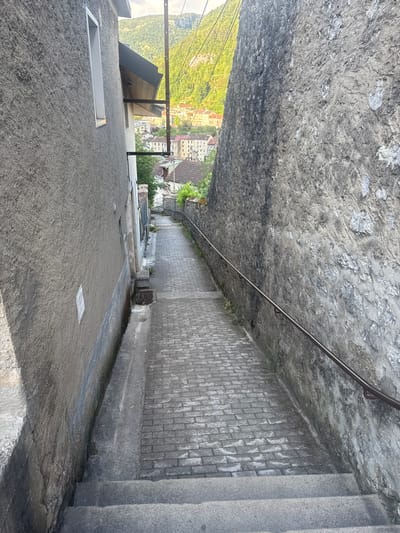 The image shows a downward view along a stone pathway and staircase in Saint-Claude, France. The pathway is bordered by a rough stone wall on the left and a brick wall on the right. A metal handrail runs along the brick wall. The pathway leads down to a street with buildings visible in the background, nestled among lush green hills. The scene appears to be set during the daytime, with bright sunlight casting shadows. The weather seems fair. No people are visible in the image. The overall impression is of an outdoor architectural setting, possibly a public walkway or a street in a historic town.