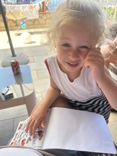 A young child with blonde hair and blue eyes is pictured indoors, leaning over a book. The child is wearing a white shirt with black stripes and a beaded necklace. They are smiling and their hand is touching their face. The book appears to be an activity book with stickers. A table is visible in the foreground, and a patio area with tiled flooring is visible in the background, suggesting an outdoor setting. The lighting suggests it is daytime. The provided location context is Sant Feliu de Guíxols, Spain.
