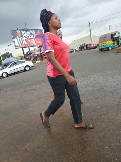A young woman is pictured from the waist down, wearing a red t-shirt with white and red patterned sleeves, dark jeans, and dark sandals. She is standing on a wet asphalt road, suggesting it may have recently rained. The scene is outdoors, with a cloudy sky overhead, indicating overcast weather.
Behind the woman, a billboard displays political campaign material. The text on the billboard reads "REDEMPTION 2027 NDC" and features a photo of a man identified as "HON JAMES YAKUBU". Another sign visible behind it has "PTA-2266" on it, which could be a vehicle registration number. To the left, a green and yellow motorized tricycle, commonly known as an "Okada" or "Keke napep" in Nigeria, is parked. Further in the background, there are other vehicles and what appear to be market stalls or shops. The setting is likely a street in a town or city in Nigeria, given the presence of the tricycle and the political billboard. The woman's gaze is directed upwards, and her expression is unreadable from this perspective.