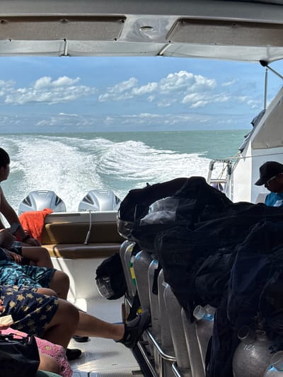 The image depicts the rear deck of a boat in motion, likely on the waters off Choeng Mon, Thailand. The foreground is occupied by several individuals seated on a cushioned bench, partially obscured by dive gear. These include diving tanks, black bags, and scuba equipment. One person, wearing a black baseball cap and sunglasses, is visible from the shoulders up, looking forward. Another person to the left is mostly obscured but appears to be wearing patterned swim trunks. The boat's twin outboard motors are visible at the stern, creating a significant wake in the turquoise water. The sky above is bright blue with scattered white clouds. The overall scene suggests a recreational boat trip, possibly for diving or snorkeling, in a tropical setting.