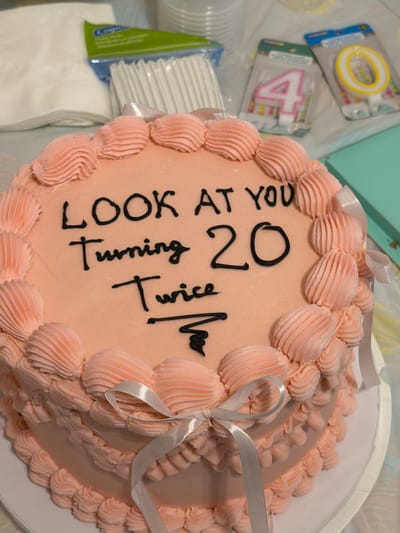The image shows a pink frosted cake with black writing that reads "LOOK AT YOU Turning 20 Twice". The cake is decorated with piped frosting shells around the top and bottom edges and a white ribbon tied around the side. In the background, there are several items on a surface, including a package of white cupcake liners, two birthday candles (a "4" and a "0"), and a stack of white napkins. A light blue box is visible in the far right background. The setting appears to be an indoor domestic environment, possibly a kitchen or dining area. There is no indication of a specific location in Saint-Césaire, Canada.