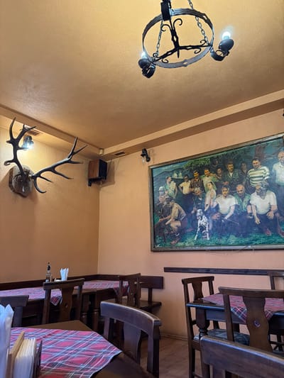 The scene depicts the interior of a dining establishment in Gaber, Bulgaria. Walls and ceiling are painted a peach-orange color, with a darker brown trim line along the upper wall perimeter. A central black wrought-iron chandelier with four exposed bulbs hangs from the ceiling. Additional lighting is provided by two wall-mounted sconces with exposed bulbs. On the left wall, a large set of deer or elk antlers is mounted on a wooden plaque. Below the trim line and to the right of the antlers, a small, dark-colored speaker cabinet is mounted. The right wall features a large, framed painting depicting a group of approximately 15-20 men, some with hunting rifles, and at least one Dalmatian dog, in an outdoor setting with distant white structures. Multiple dark wooden tables, each covered with a red and white plaid tablecloth, are arranged throughout the room. Seating includes dark wooden chairs with carved backs and built-in wooden bench seating along the walls. A table in the foreground displays a napkin dispenser and two small bottles. The floor is wooden. No individuals are present in the depicted environment.