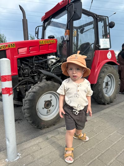 A young child stands beside a red Belarus 320.4M tractor in an outdoor setting, likely in the city of Odintsovo, Russia. The child, wearing a beige hat with animal features, a light-colored shirt patterned with bear images, dark shorts, and brown sandals, is positioned between the tractor's front tire and a white pole marked with red tape. The tractor is a Belarus model, as indicated by the Cyrillic script on its side. The environment is paved with gray brick. The sky appears overcast.