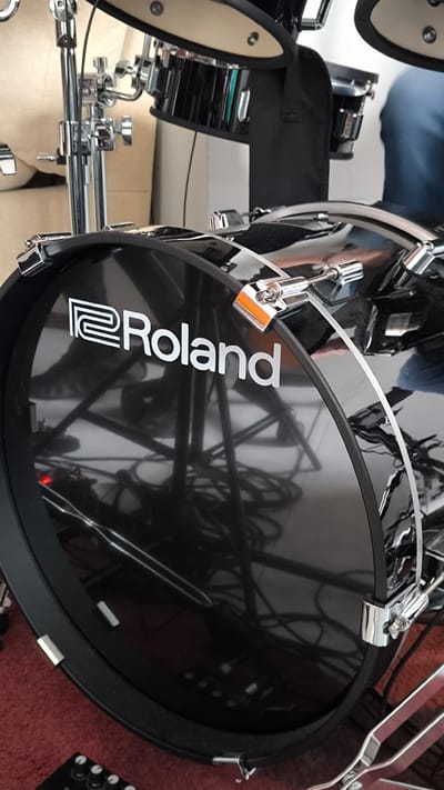 The image shows a close-up of a Roland electronic drum set. The prominent feature is the black bass drum with the white "Roland" logo. Drum hardware, including tension rods and mounting brackets, are visible. Behind the bass drum, parts of other drum components and stands are partially visible. Cables and wires are visible on the floor. The environment consists of a carpeted floor of an indistinguishable color. The image does not give any context about the location in Ripollet, Spain. There are no people depicted in the image.